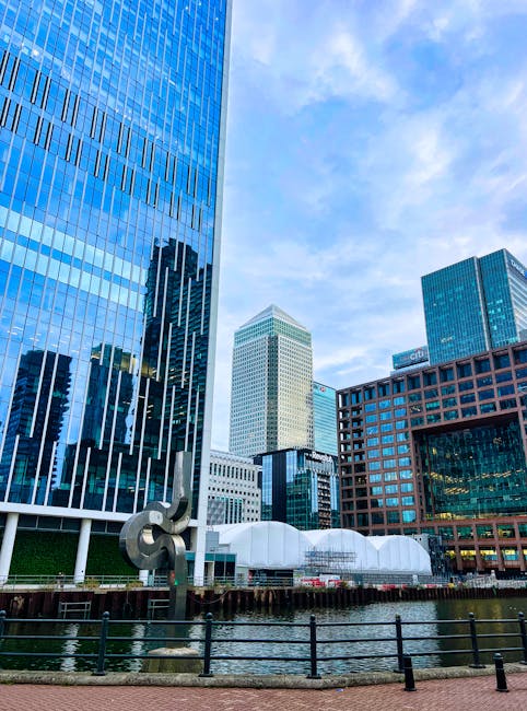 A cityscape featuring modern high-rise buildings with extensive glass facades reflecting the sky and surrounding structures. In the foreground, a canal runs parallel to the buildings, bordered by a bl
