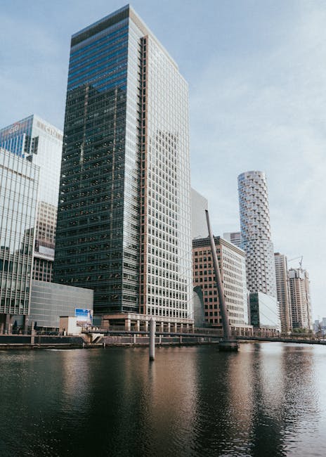 A cityscape featuring modern high-rise buildings with extensive glass facades reflecting the sky and surrounding structures. In the foreground, a canal runs parallel to the buildings, bordered by a bl